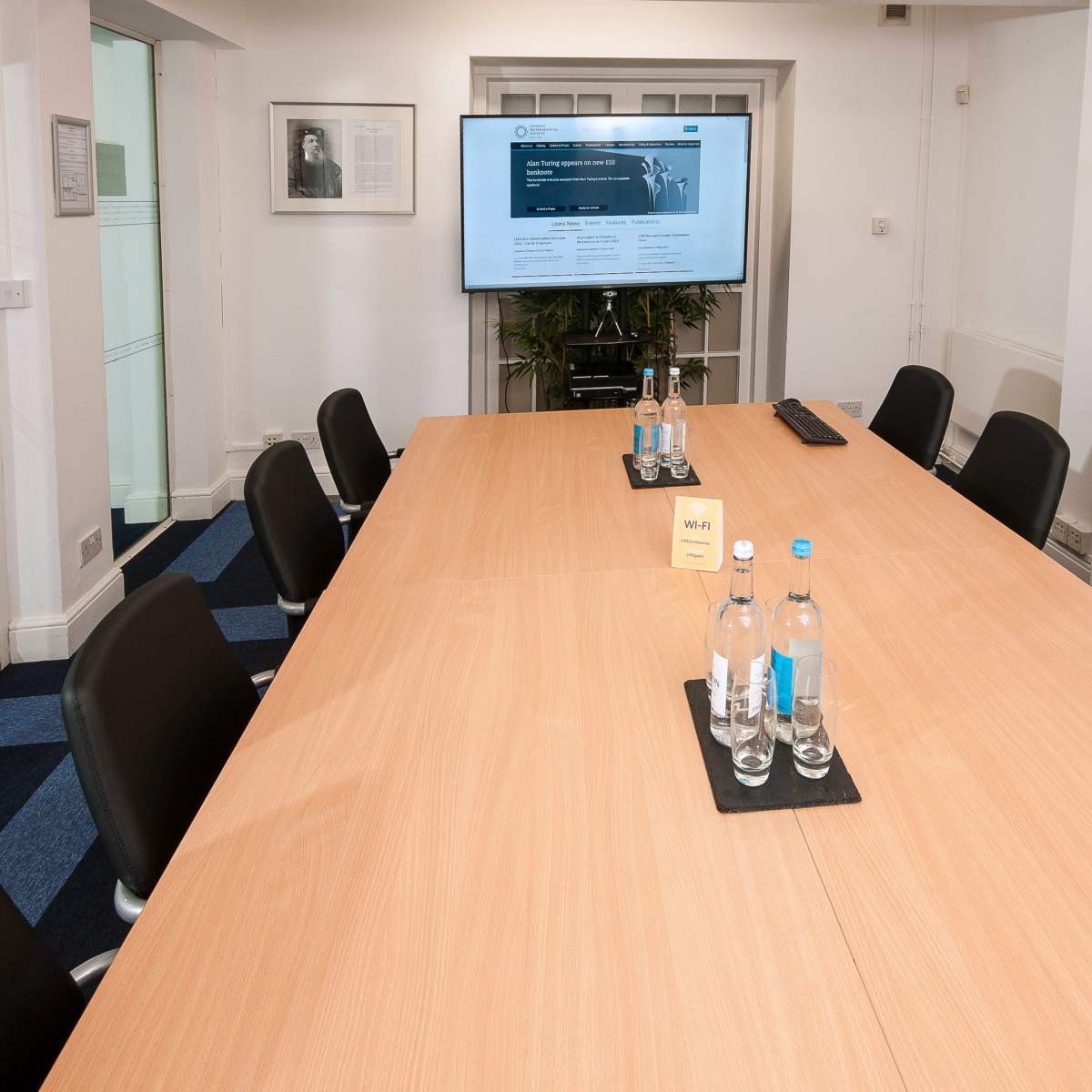 A modern conference room features a large wooden table surrounded by black chairs, with a screen displaying a website in the background.