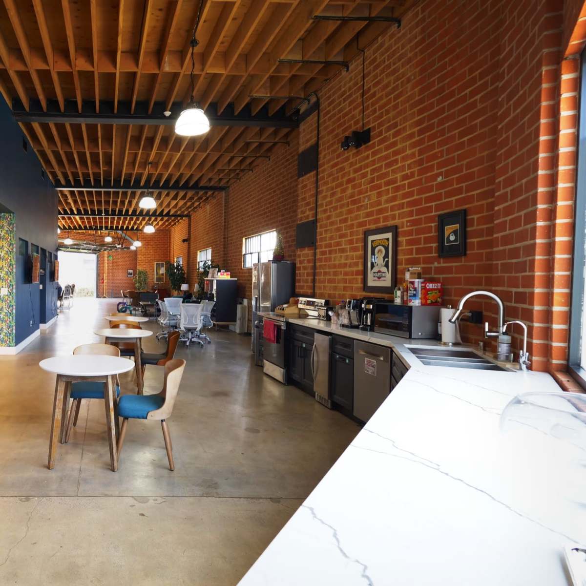 A spacious, industrial-style interior with exposed brick walls, wooden beams, a dining area featuring a round table, and a kitchen along one side.