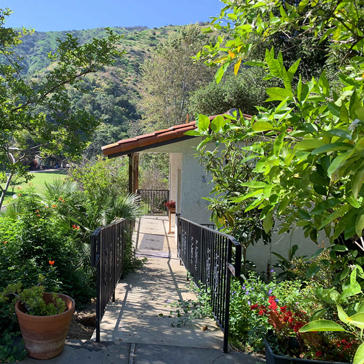 A pathway leads through lush greenery to a house with a terracotta roof, set against a hillside.