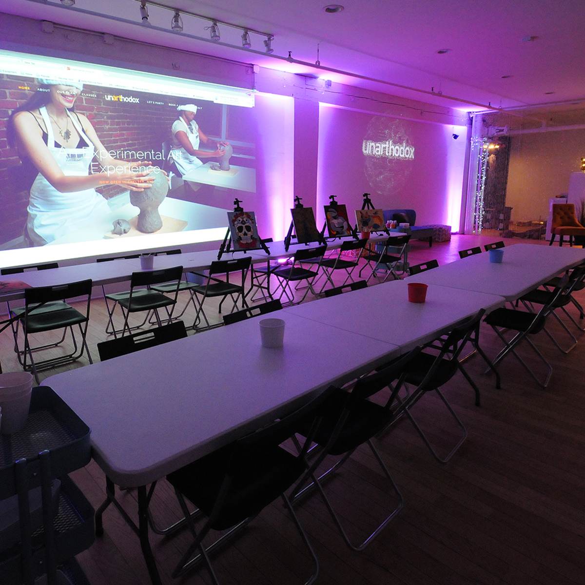 A spacious room is set up with a long table and chairs, featuring a large projector screen displaying a website, illuminated by purple lights.