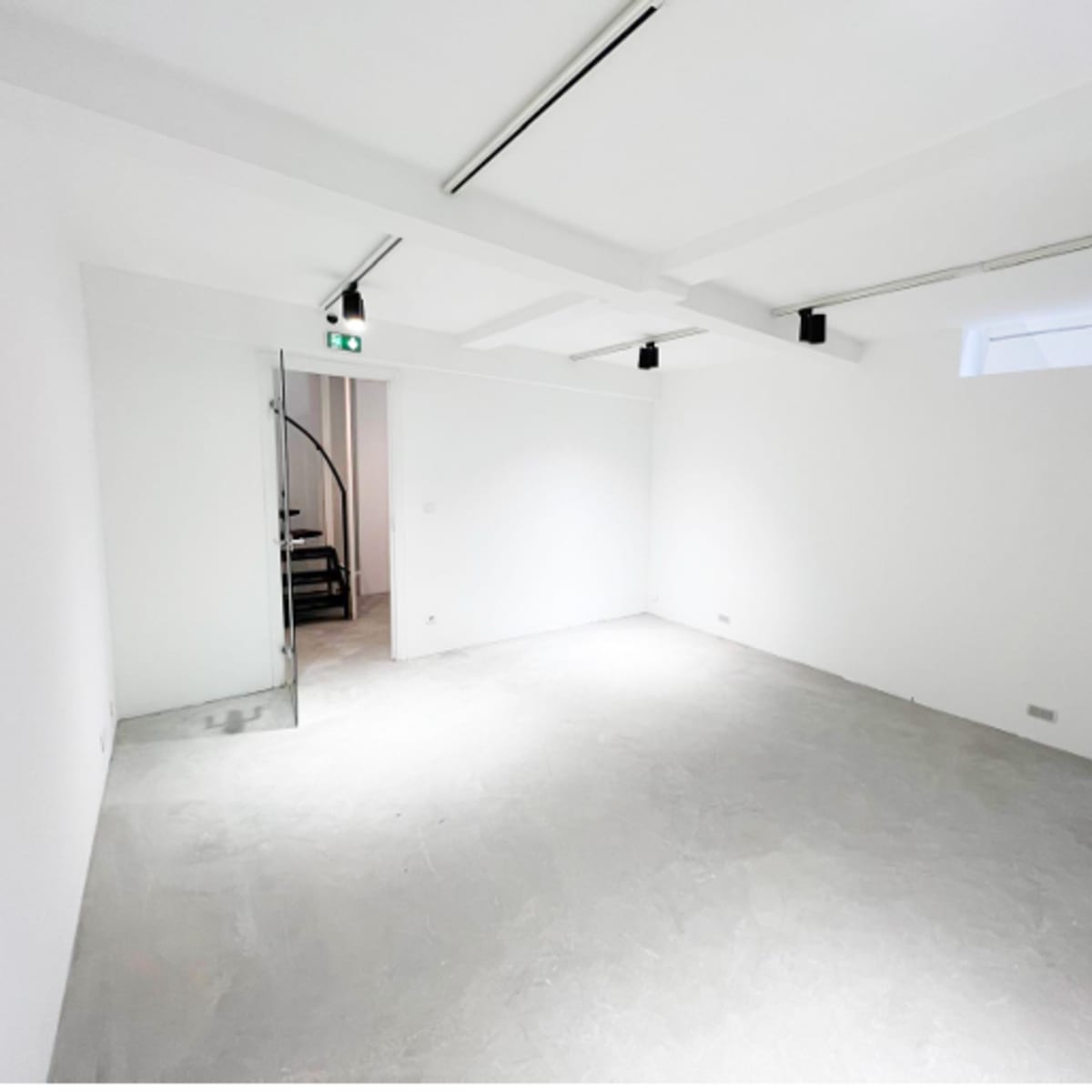 A minimalistic, empty room with white walls, a concrete floor, and a staircase visible in the background.