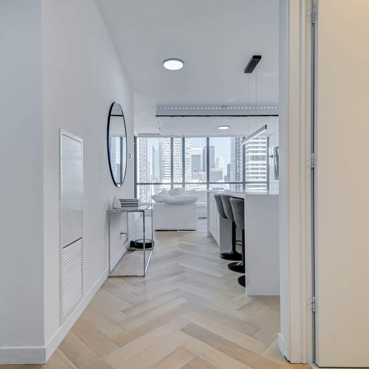 A hallway with herringbone wooden flooring leads to a bright living area featuring large windows and modern furnishings.