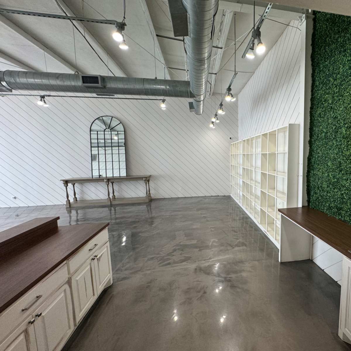 A spacious room with a concrete floor, a green vertical wall, shelving units, a sink, and overhead lights.