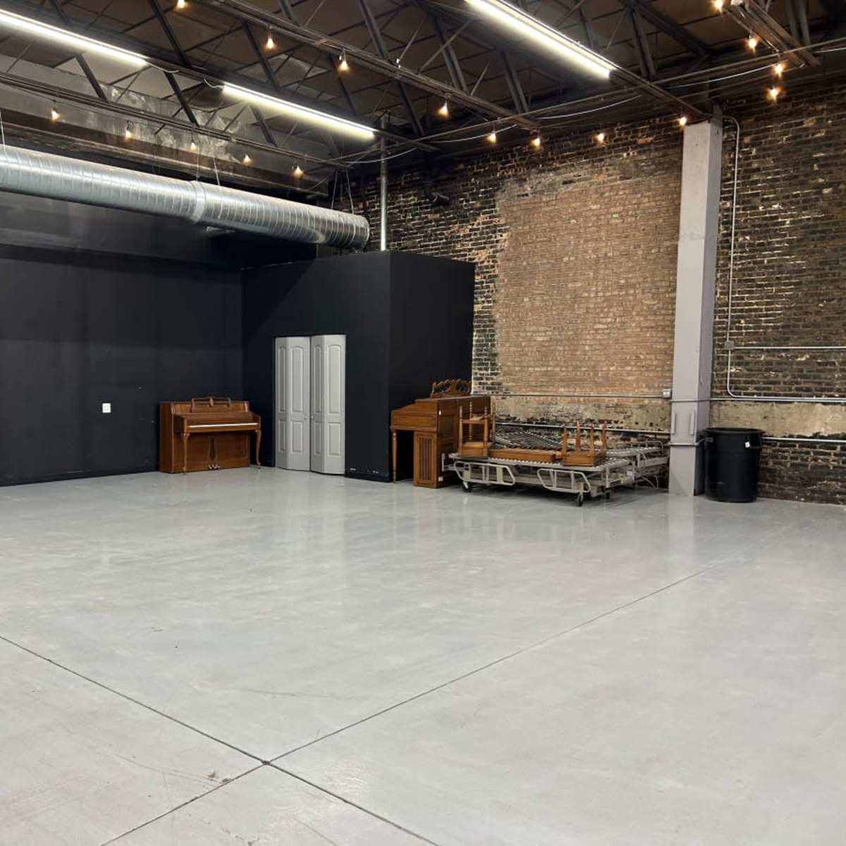 An empty warehouse space with gray concrete floors, exposed brick walls, and various pieces of furniture against one wall.