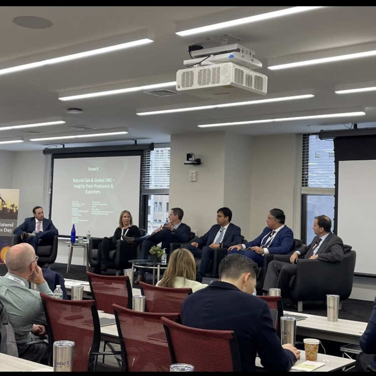 A panel discussion is taking place in a conference room, featuring six speakers seated on a stage with a presentation displayed on the screen behind them.