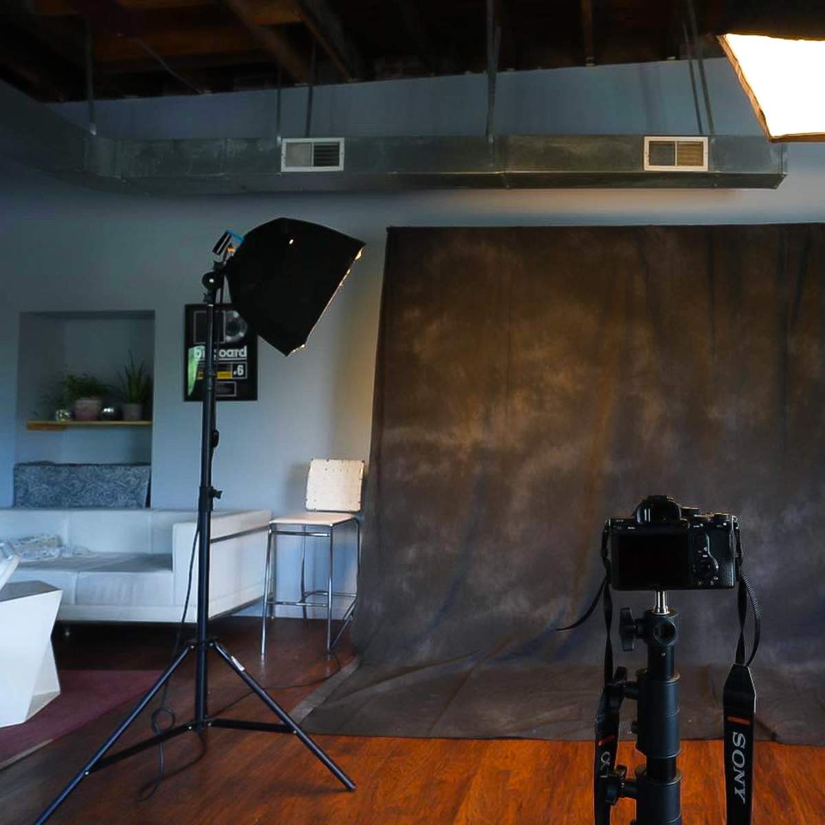 A photography studio setup with a camera on a tripod, a dark backdrop, lighting equipment, and a few clothing items hanging nearby.