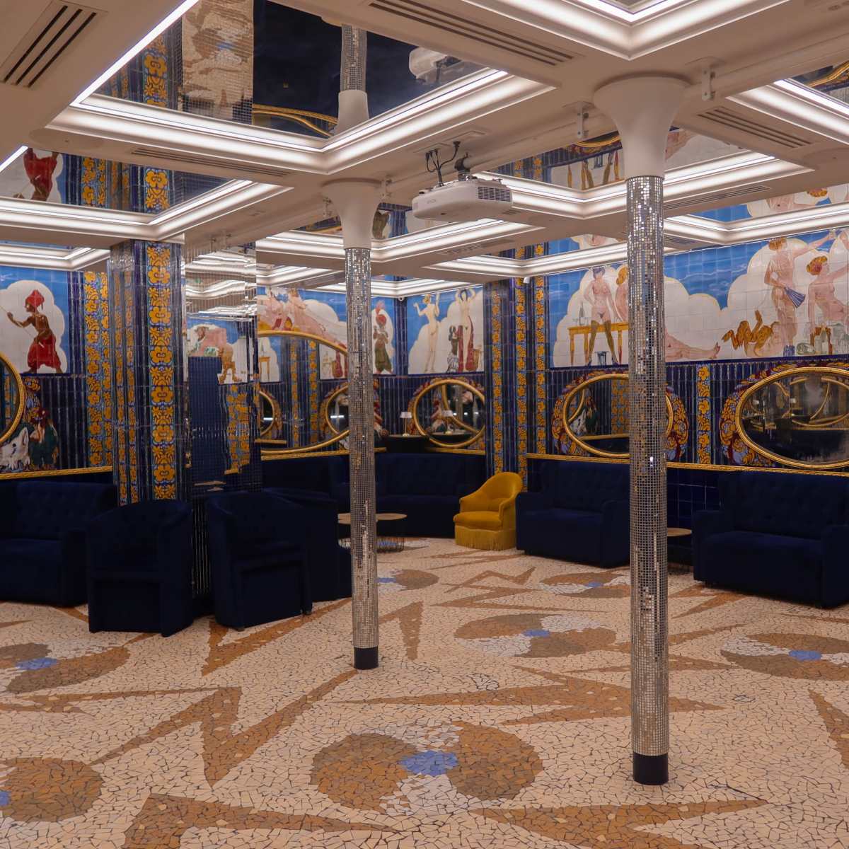 A brightly lit lounge area with blue velvet sofas, circular mirrors on the walls, and intricate tiled flooring featuring geometric patterns.