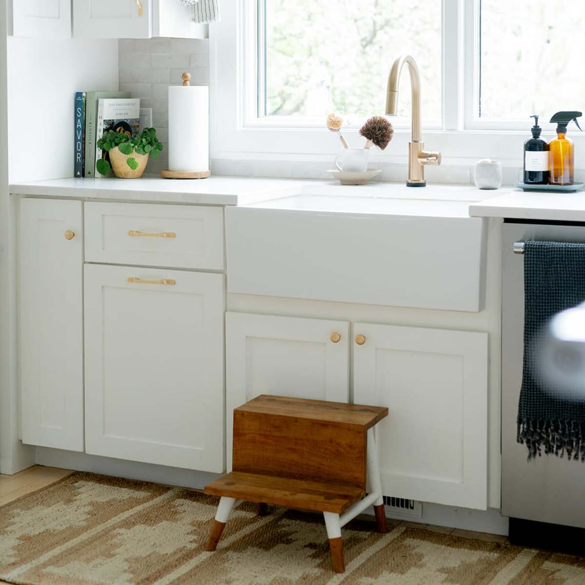 A bright kitchen features white cabinetry, a farmhouse sink, a wooden step stool, and a window with a view of greenery outside.