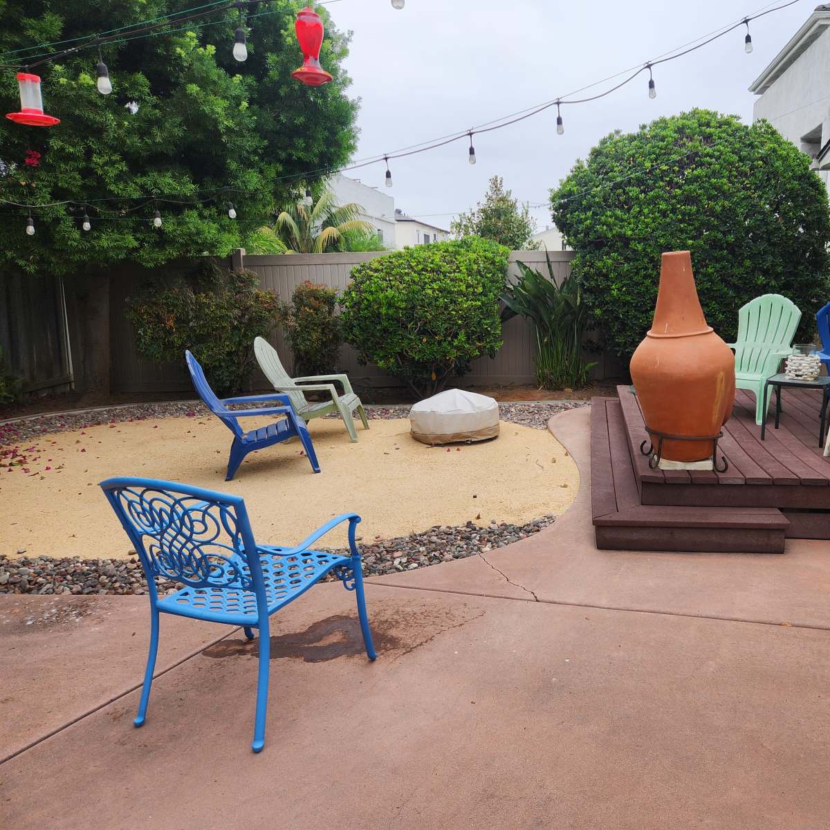 The backyard features a sand area, several chairs, a large terracotta pot, and a wooden deck beside a house.