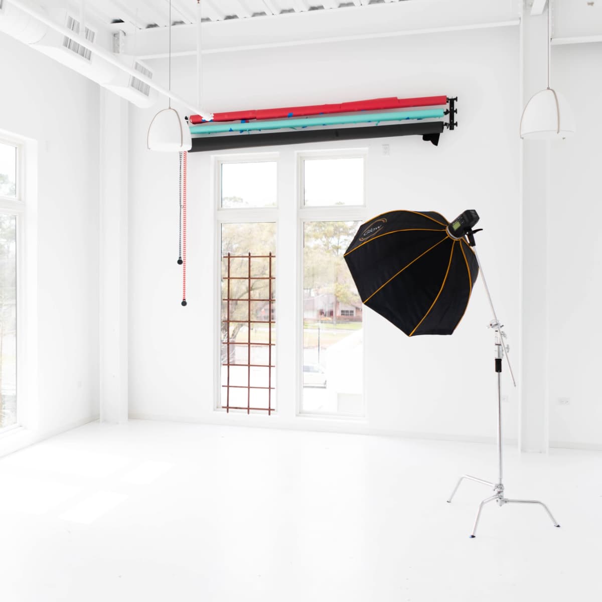 The interior of a bright studio features large windows, a mirror, and a black umbrella light stand with colorful backdrop poles hanging from the ceiling.