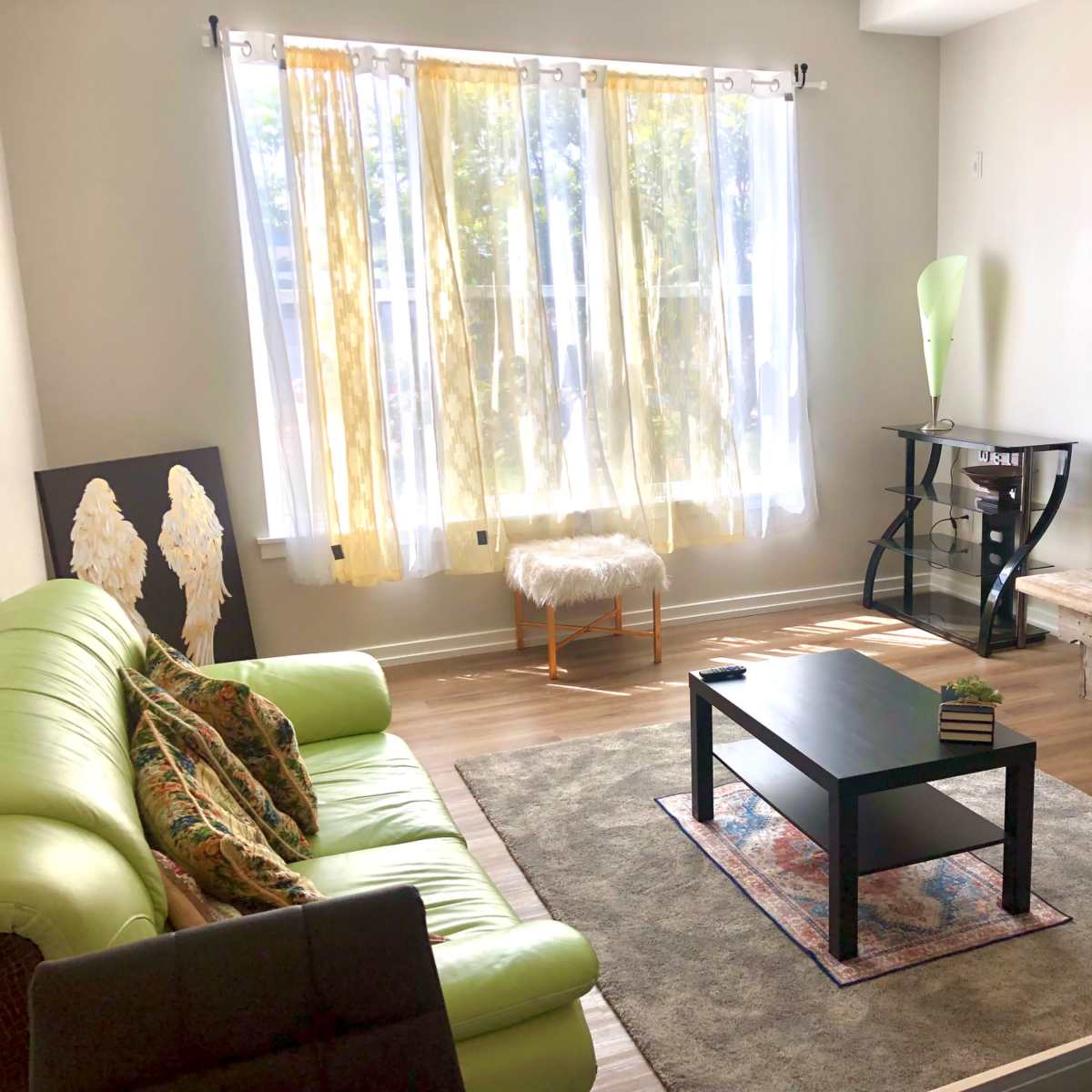 A bright living room with a green sofa, a black coffee table, and a television on a wooden stand, complemented by decorative elements and large windows with yellow curtains.