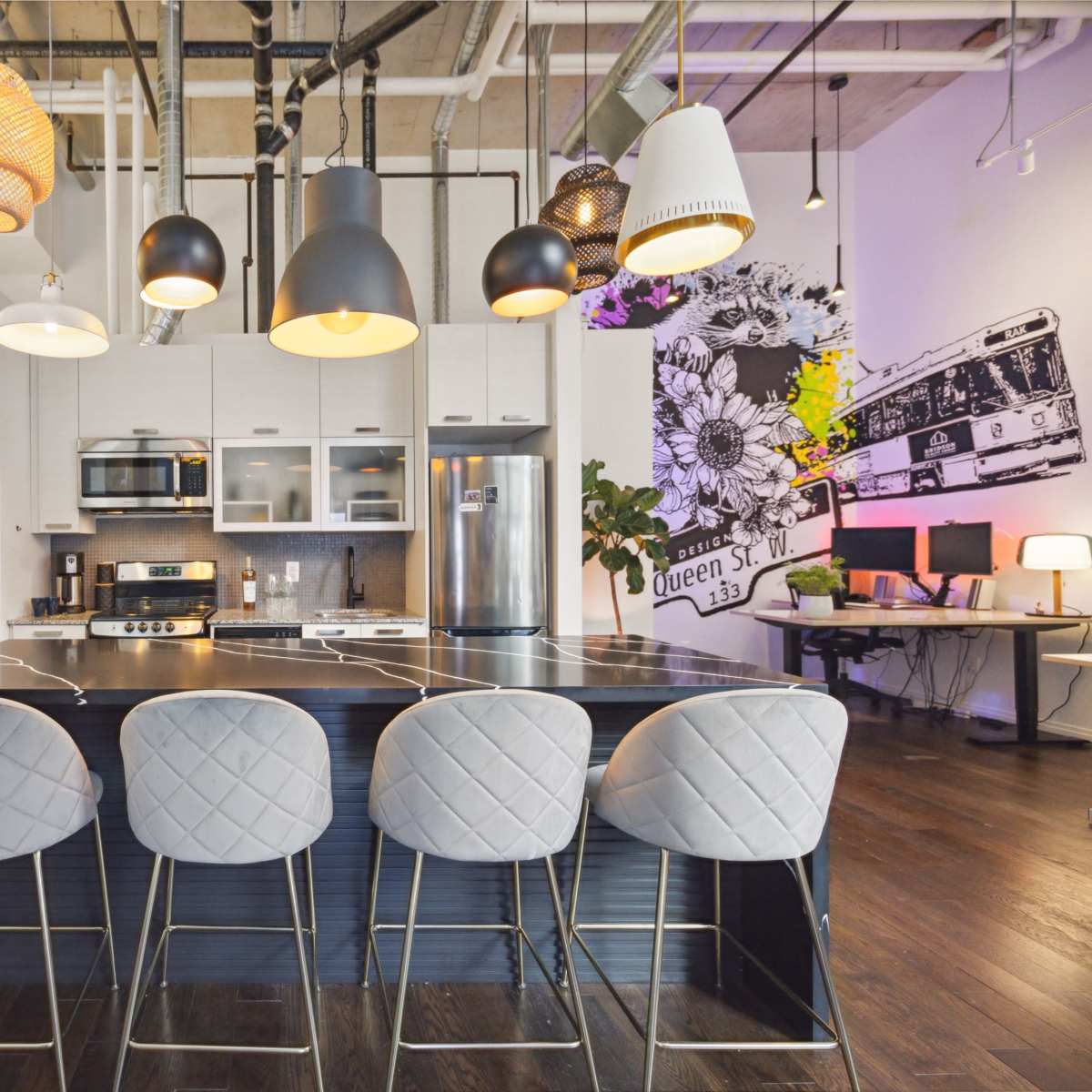 A modern kitchen features a central island with four barstools, pendant lighting, and a mix of appliances against a backdrop of vibrant wall art.