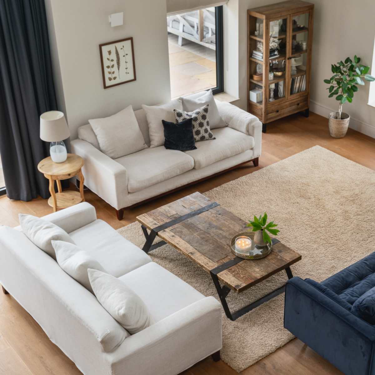 A cozy living room with three sofas arranged around a wooden coffee table on a large area rug, flanked by windows and a plant.