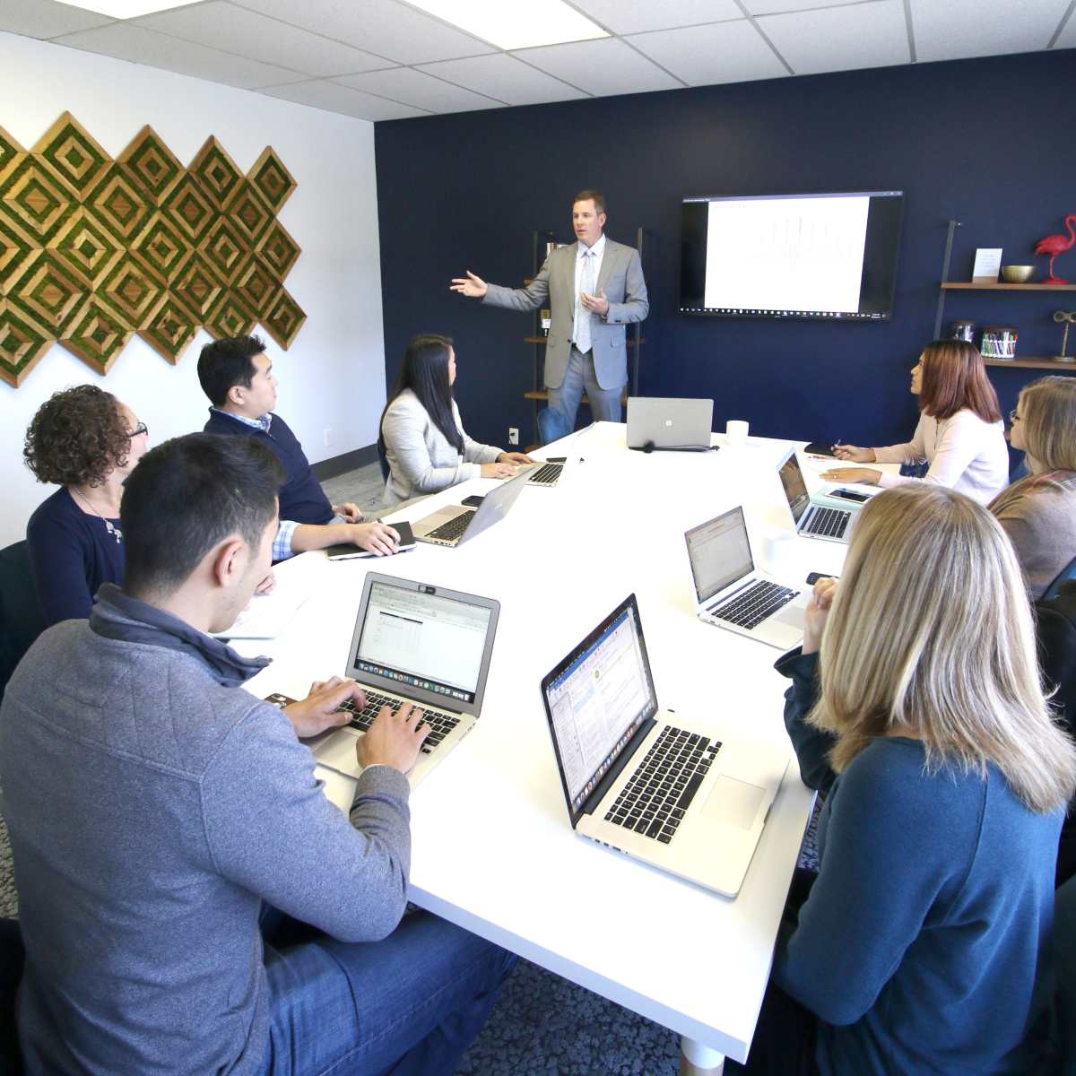 A group of eight professionals is engaged in a meeting around a large table, with one person presenting in front of a screen displaying data.
