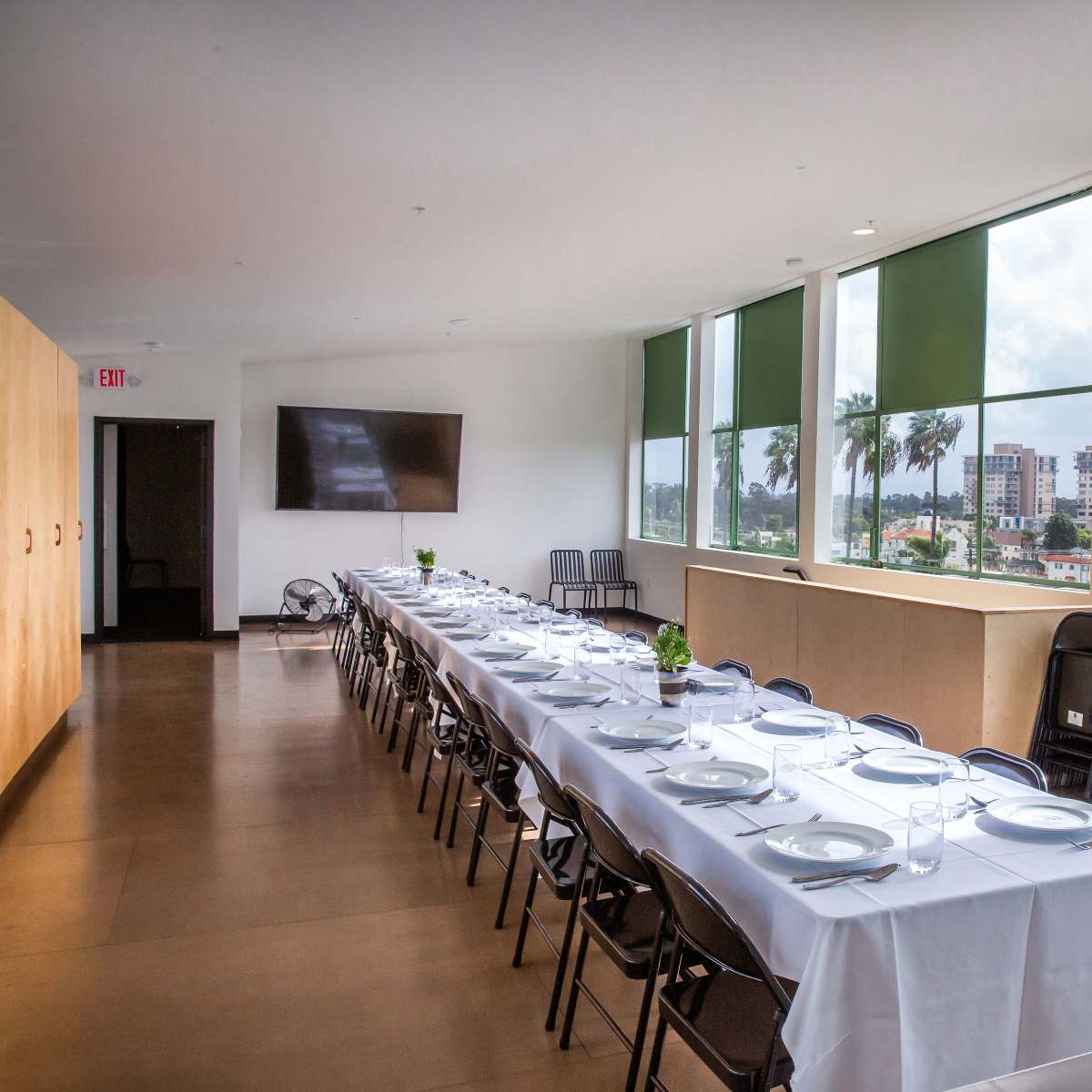 A long dining table is set with plates and glasses in a bright, modern room featuring large windows that overlook a cityscape.