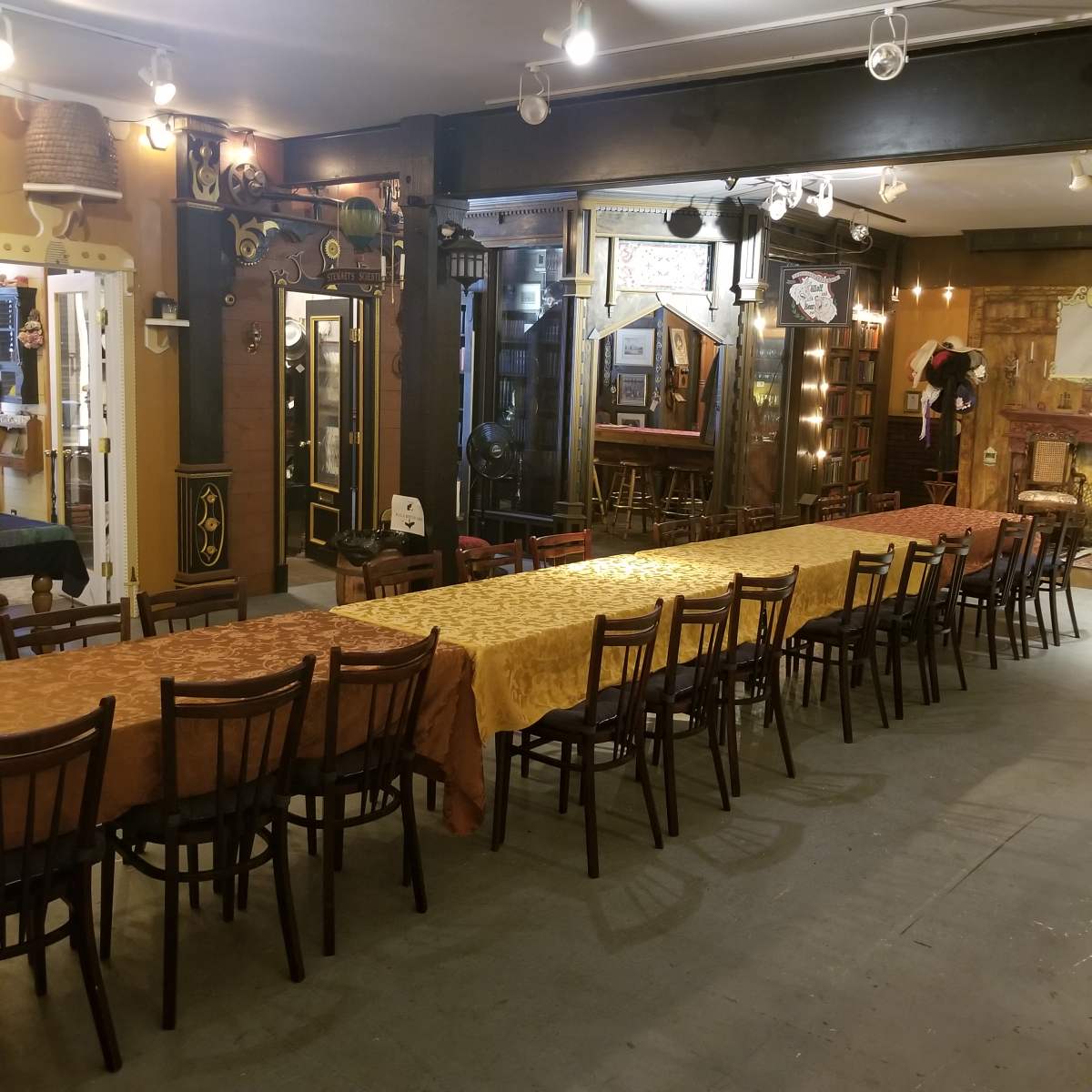 A long table set with orange and gold tablecloths, surrounded by wooden chairs, in a room with a vintage decor and various furnishings along the walls.