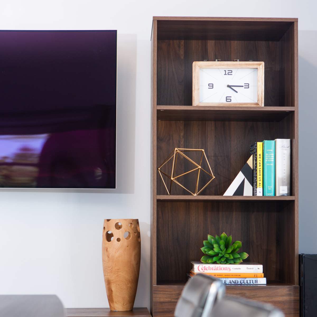 A wooden shelf displays a clock, a decorative geometric shape, books, a potted plant, and a carved vase beside a wall-mounted television.