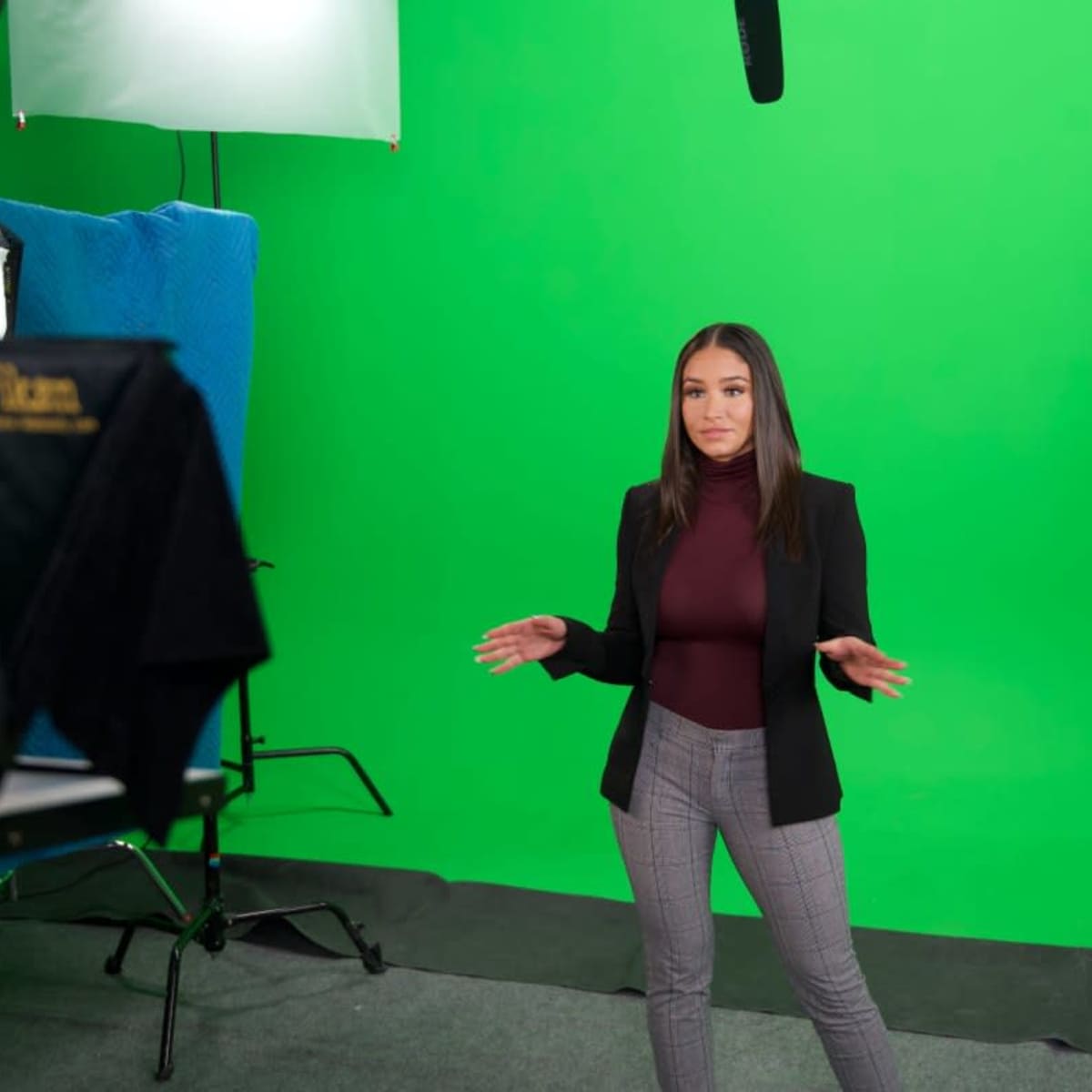 A person in professional attire is standing in front of a green screen setup, gesturing with their hands.