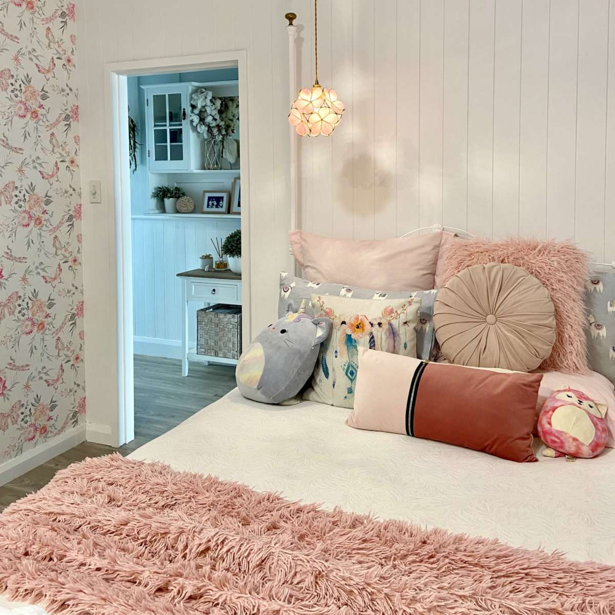 A cozy bedroom features a bed with decorative pillows and a pendant light, set against floral wallpaper and an open doorway leading to another room.