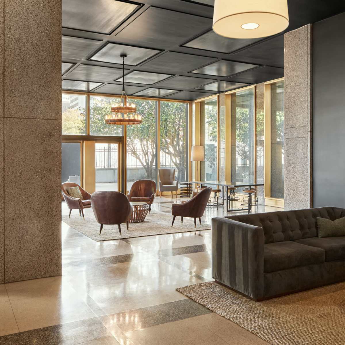 A modern lounge area with a gray couch, brown leather ottoman, and multiple seating options arranged around large windows.