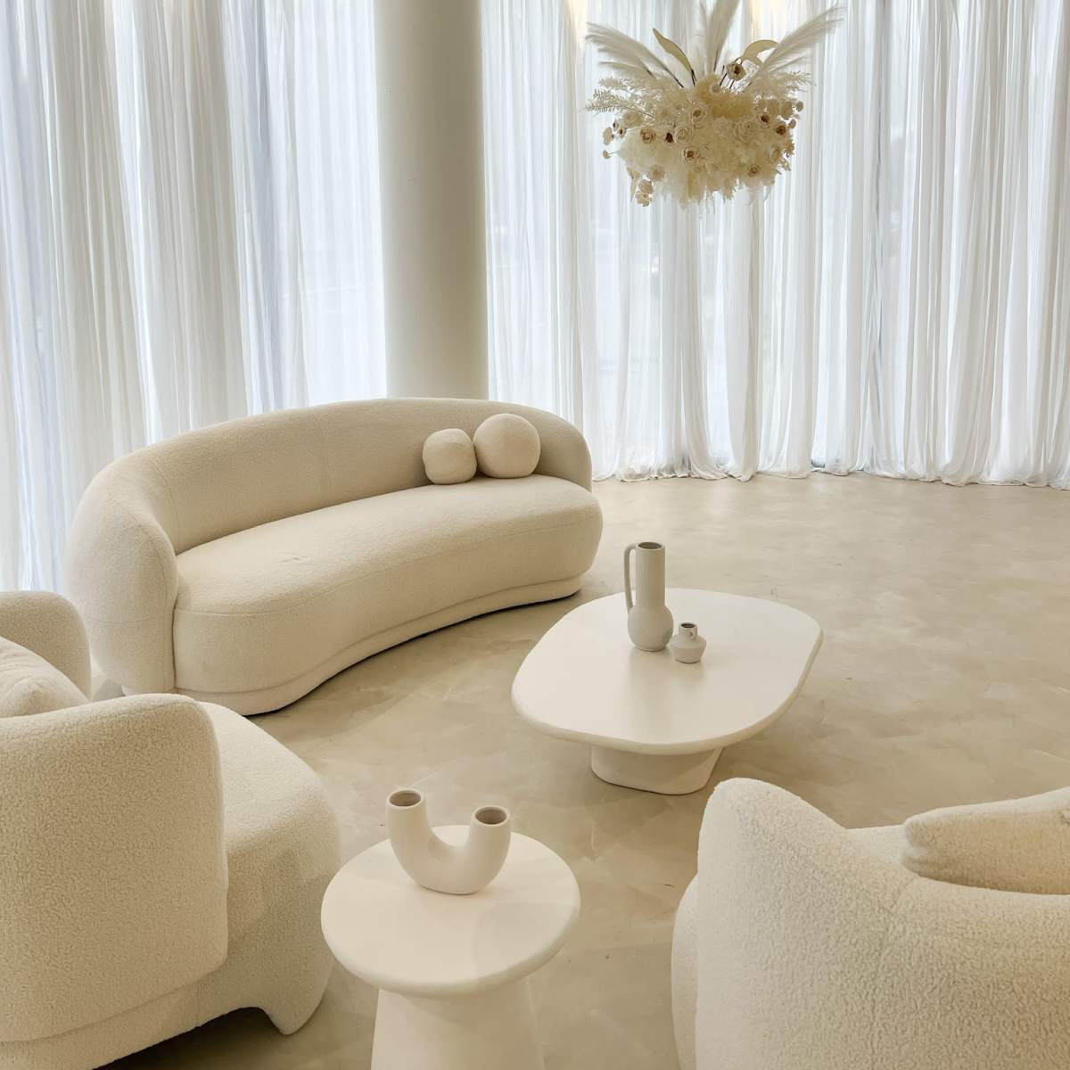 A modern, minimalist living space features white, curved furniture and sheer curtains, with a decorative light fixture overhead.