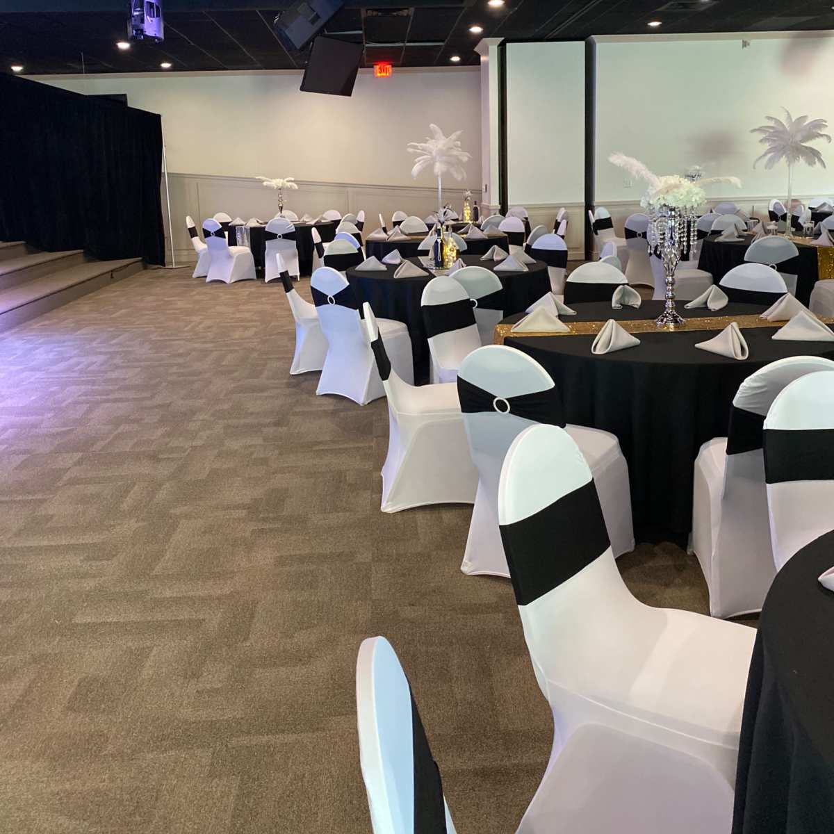 A banquet hall set up with round tables covered in black and white tablecloths, surrounded by chairs draped in white fabric with black accents.