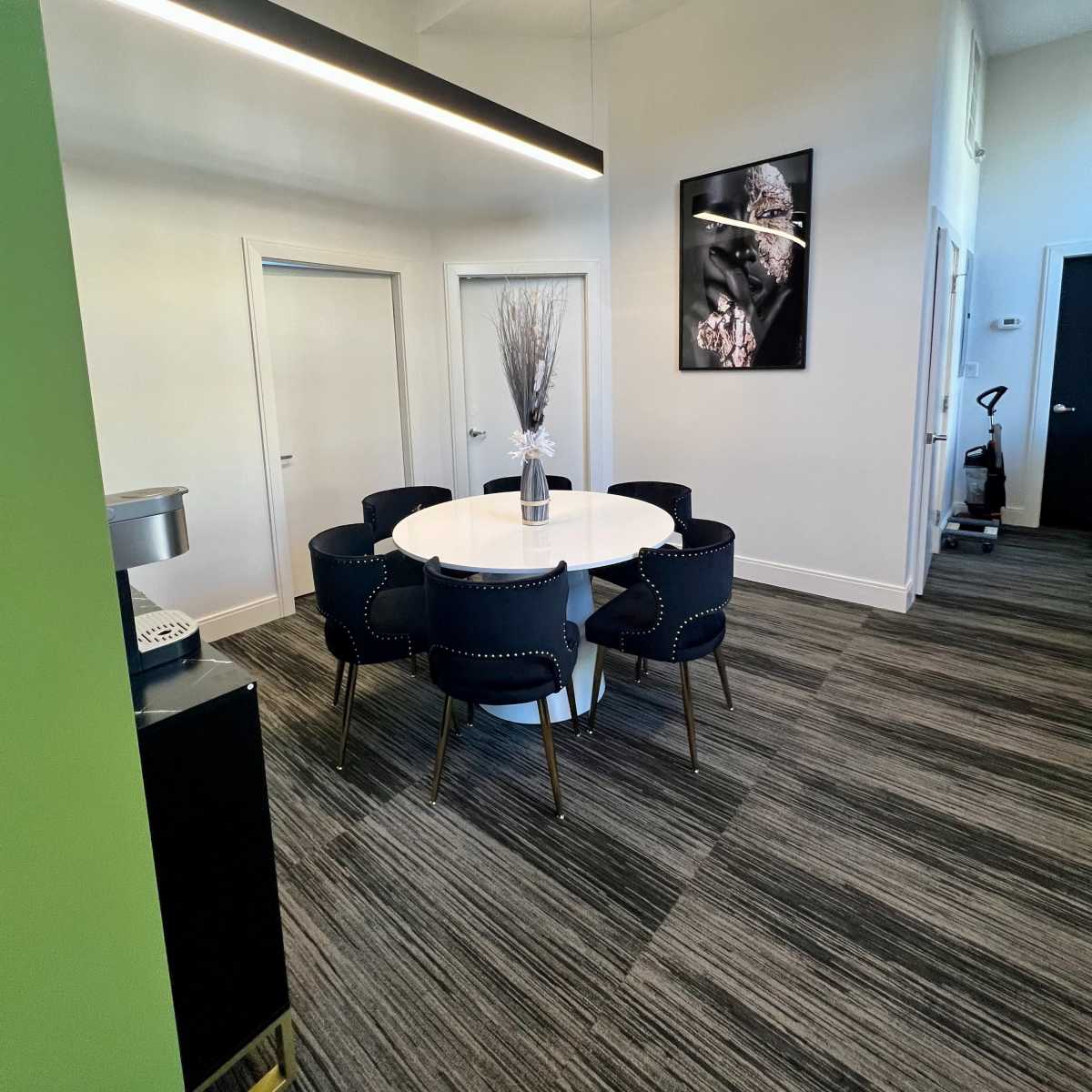 A modern office space featuring a round table surrounded by six chairs, with a vase of branches on the table and a large framed artwork on the wall.