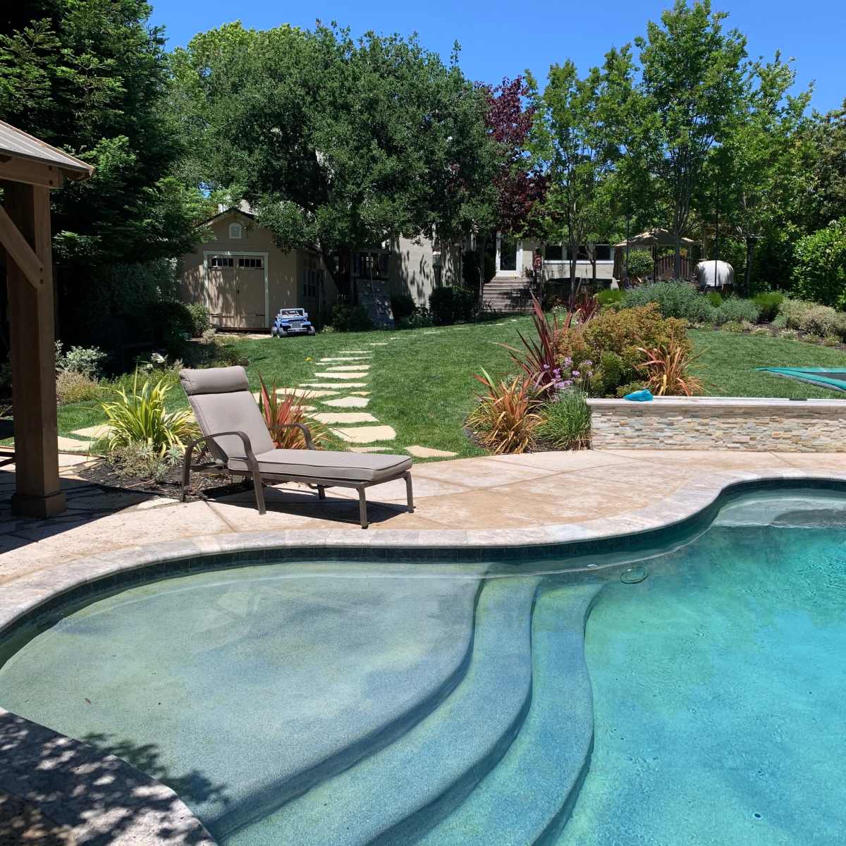 A swimming pool is situated in a backyard with a stone patio, surrounded by landscaping and a grassy area leading to a pathway and a house in the background.