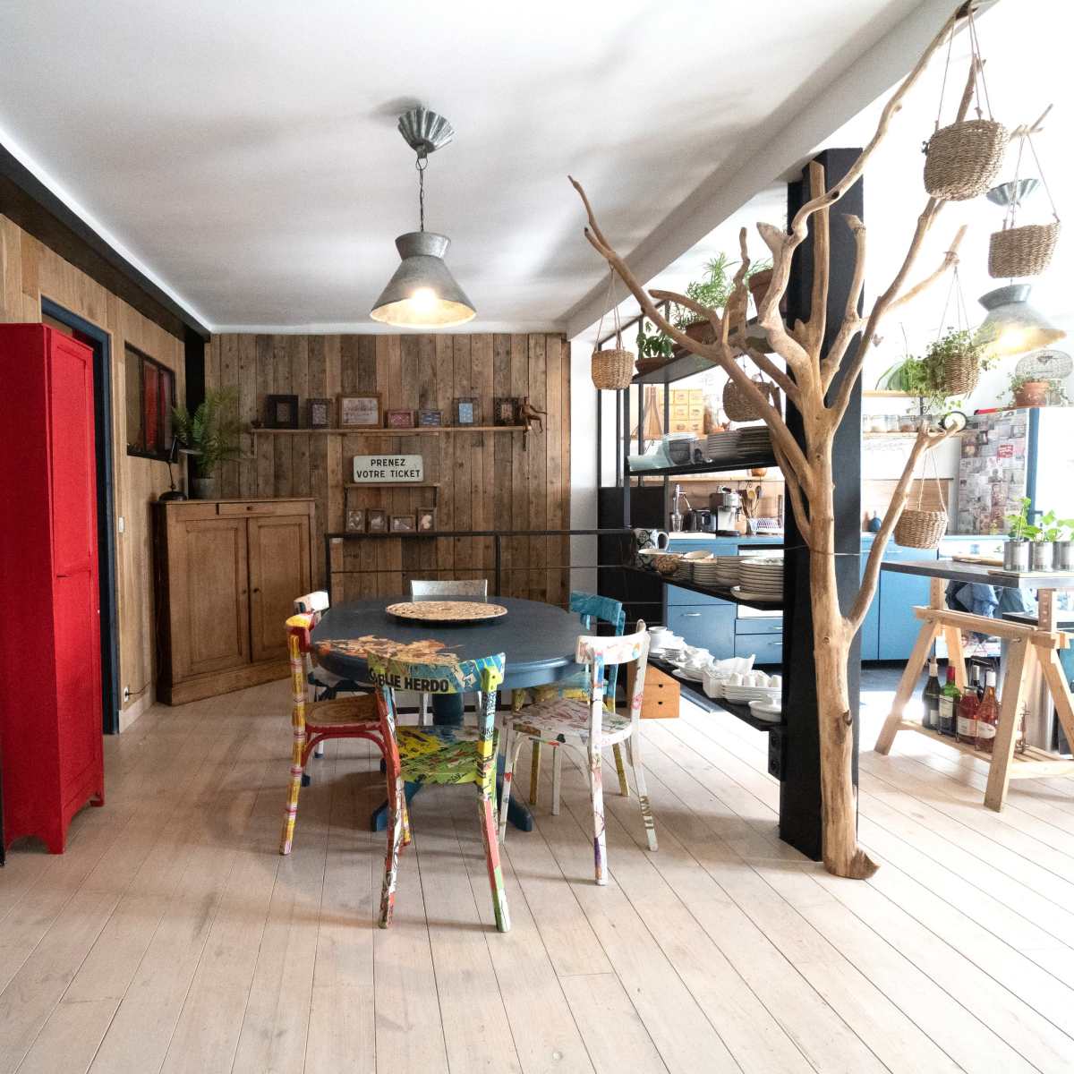 A cozy interior space featuring a dining table surrounded by colorful chairs, a wooden bookshelf, and various plants integrated into the rustic decor.