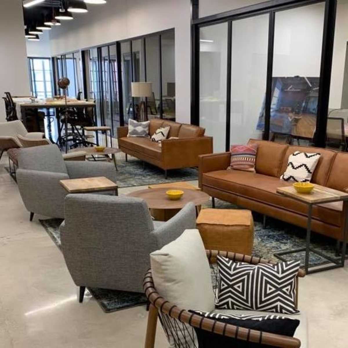 A modern office space featuring a seating area with brown leather sofas, gray armchairs, and decorative cushions, surrounded by glass-enclosed meeting rooms.