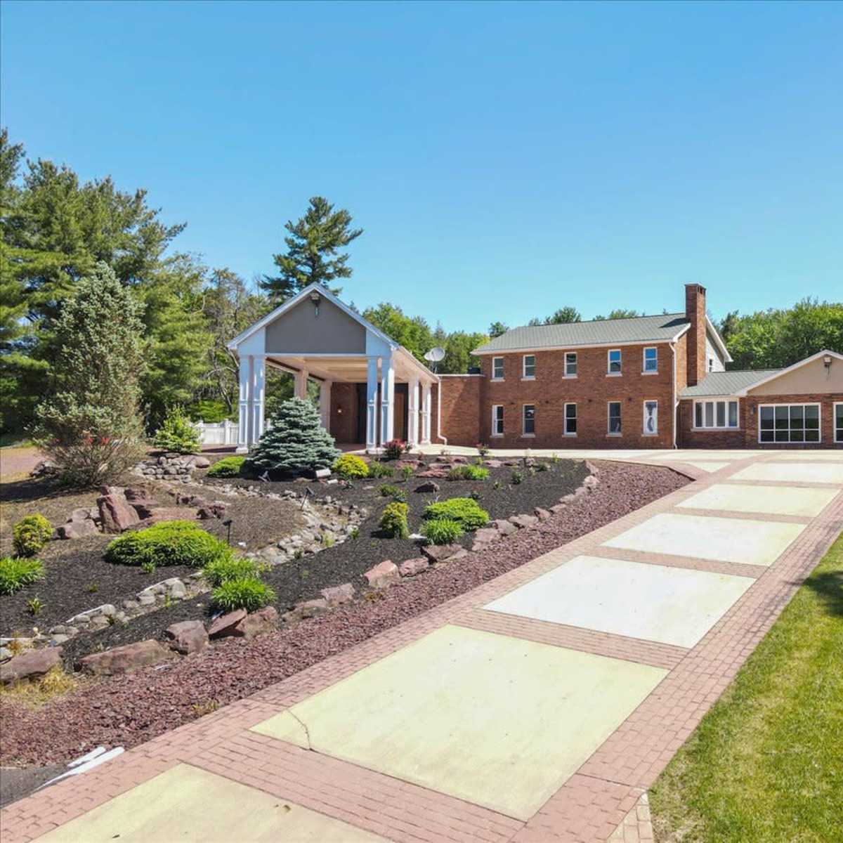 A large brick house with a white columned entrance, surrounded by landscaped gardens and a paved driveway leading to a garage.
