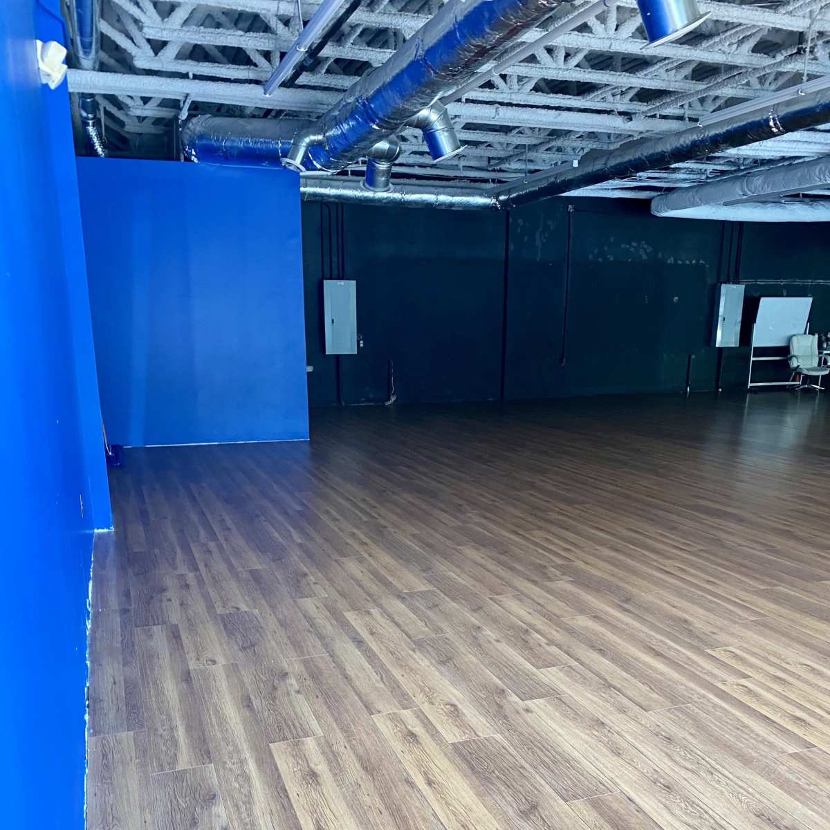 A spacious room with wooden flooring, one blue wall, and a dark wall opposite it, along with ceiling-mounted pipes.