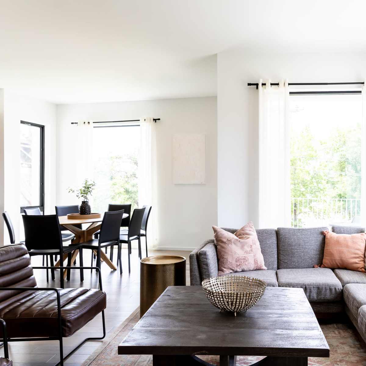 A bright living room with a gray sectional sofa, a dark wooden coffee table, and a dining area featuring a round table and black chairs.