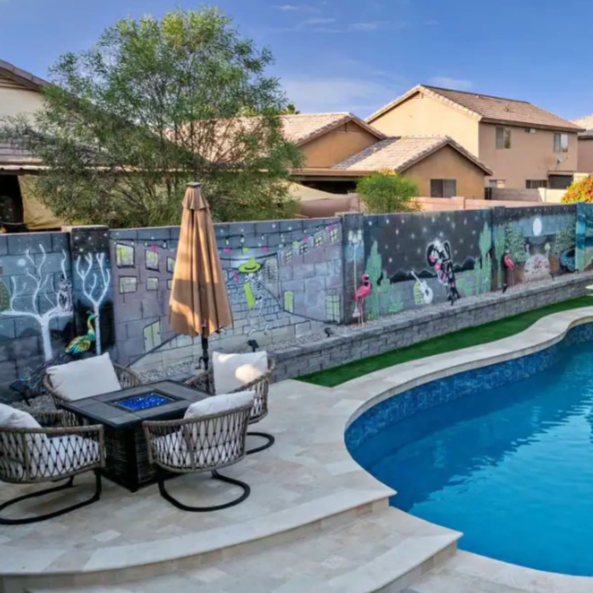A backyard features a swimming pool surrounded by lounge furniture and a mural on the wall.