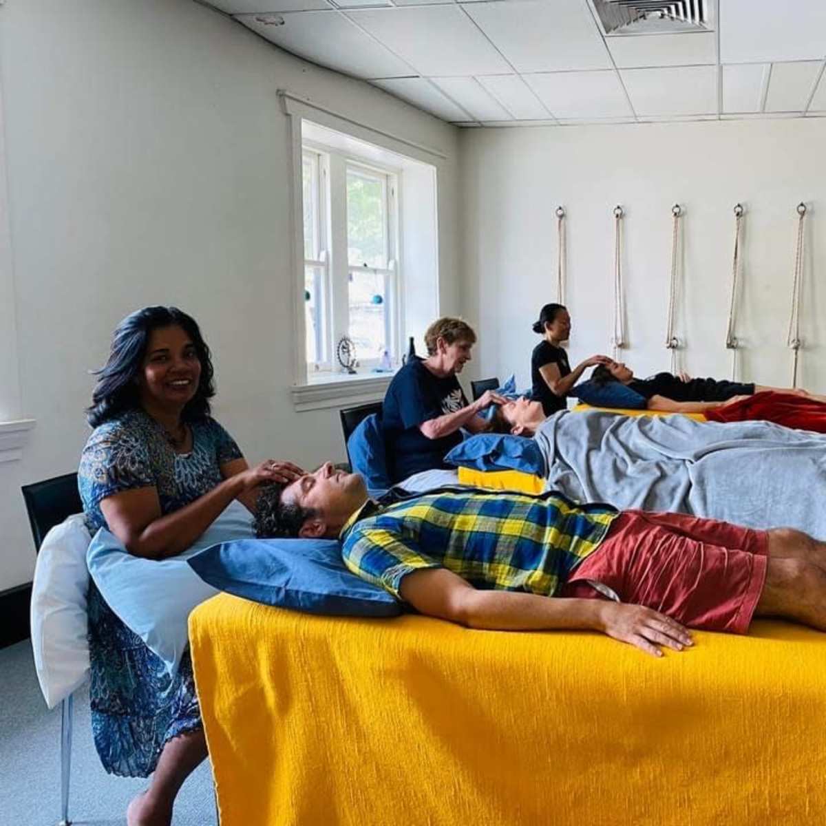 A group of five people is engaged in relaxation activities in a room, with two individuals receiving massage on treatment tables while others sit nearby.