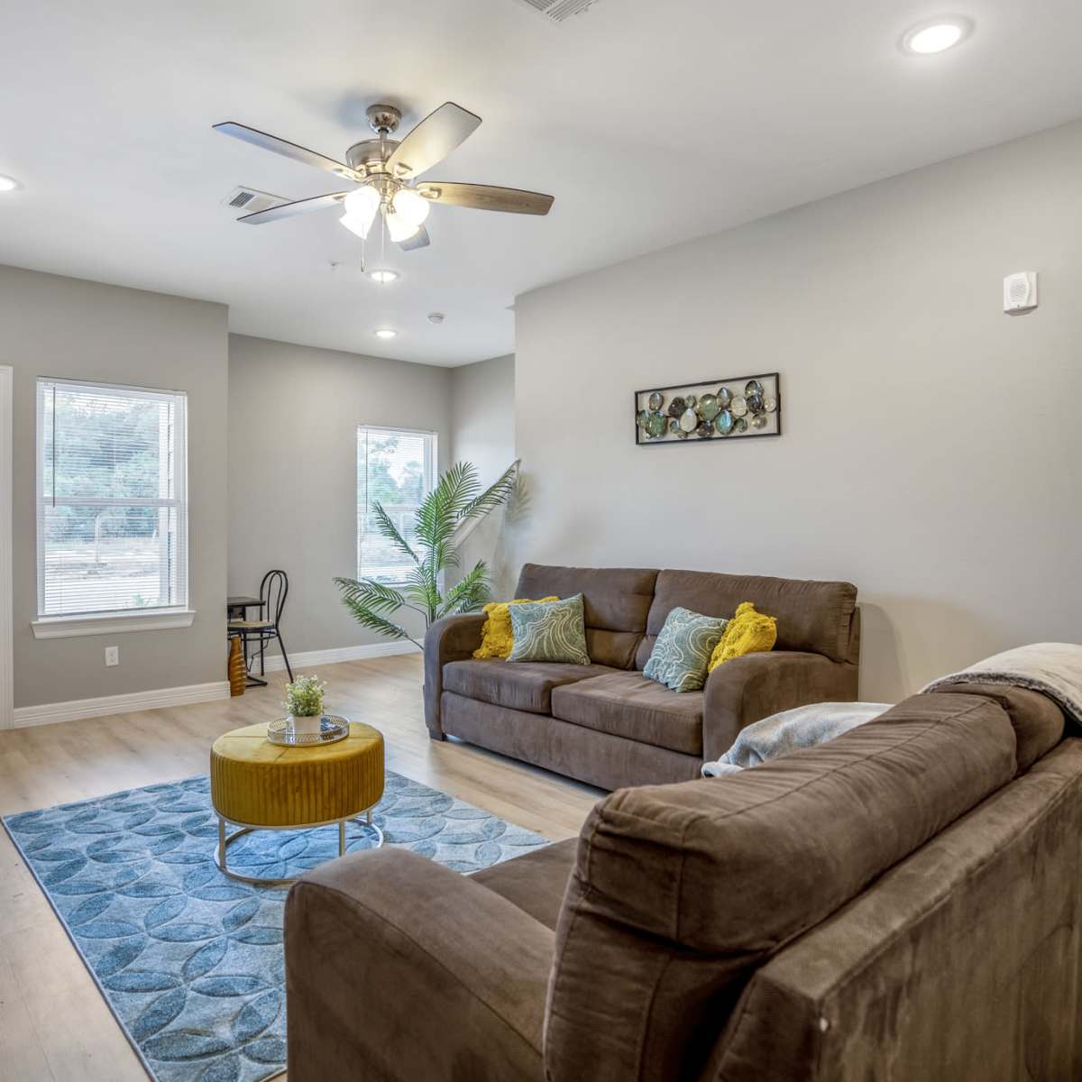 A living room features a brown sofa and armchair, a coffee table with a plant, and a window with natural light.