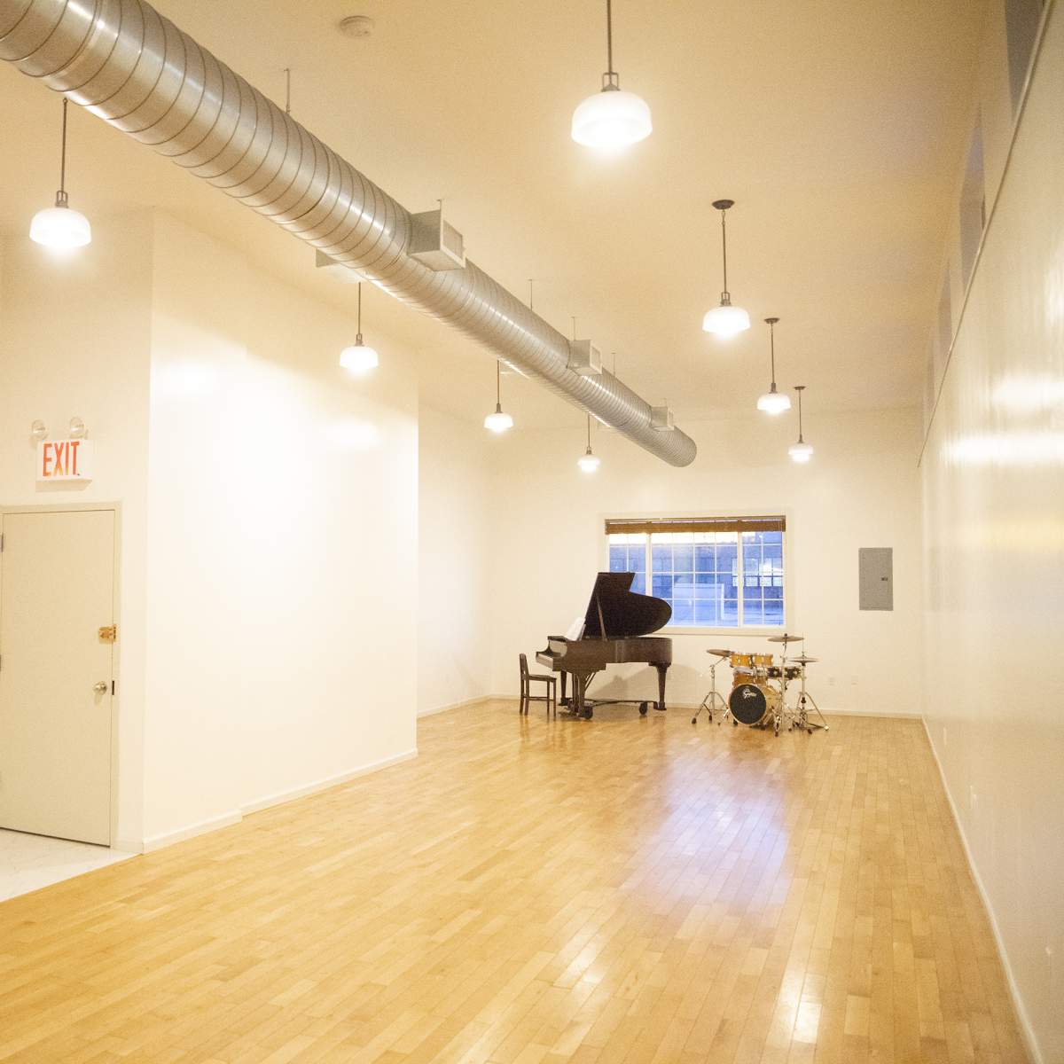 A spacious, well-lit music studio featuring a grand piano and a drum set against a white wall.