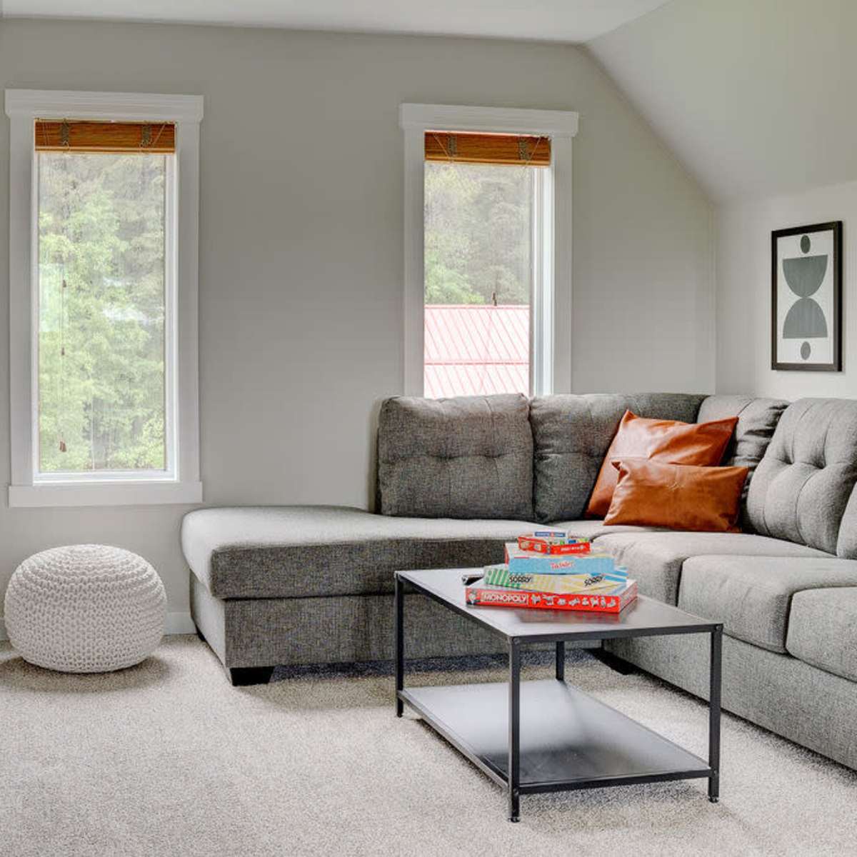 A modern living space features a gray sectional sofa, a coffee table with books, and two poufs, with large windows allowing natural light to enter.