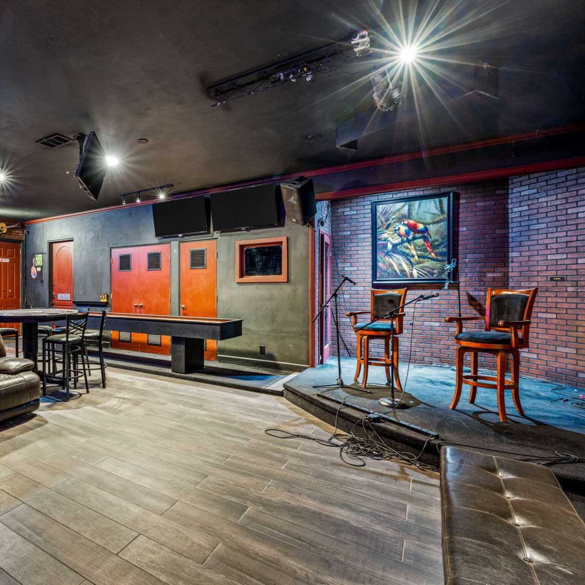 An indoor performance space featuring a small stage with two chairs, a bar area, and lounge seating arranged in a room with wooden flooring and a brick wall.