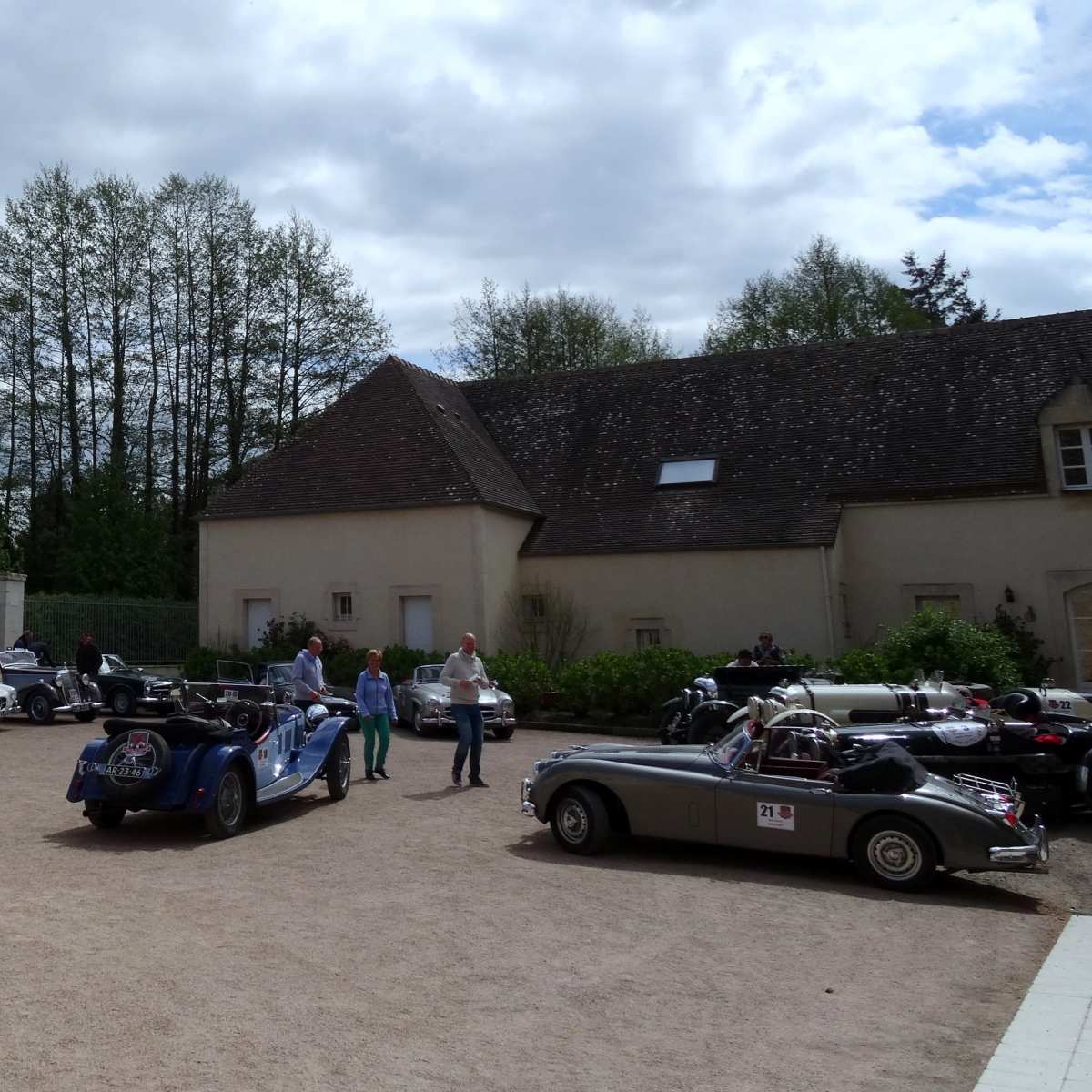 A gathering of vintage cars is set in a courtyard next to a stone building surrounded by trees under a partly cloudy sky.
