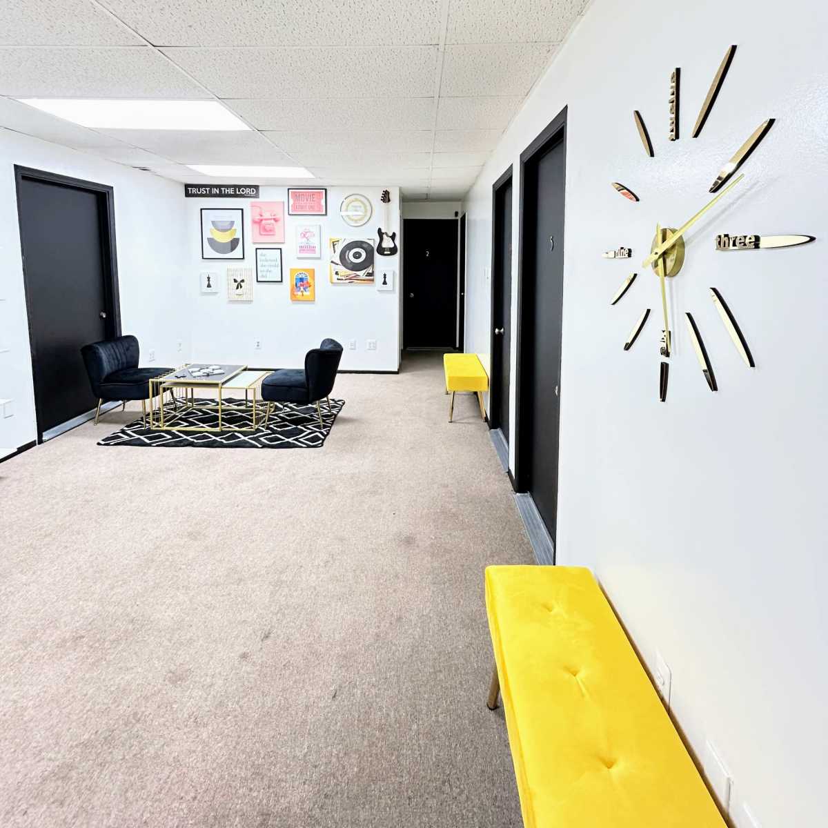 A hallway with yellow benches, black doors, a patterned rug, and a wall decorated with framed art and a large clock.