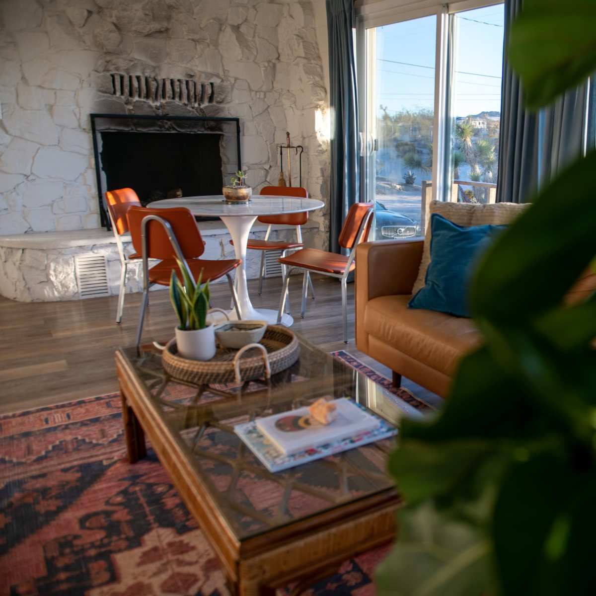 A cozy living room features a stone fireplace, a wooden coffee table with plants and magazines, and a dining area with a round table and orange chairs.