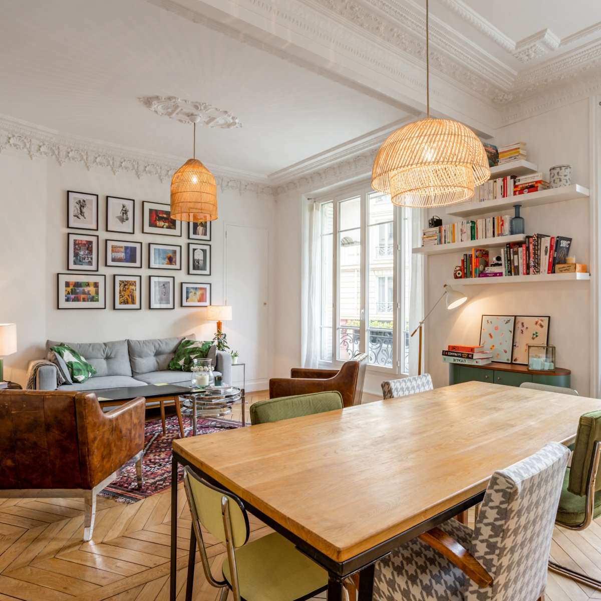 A stylish living area featuring a dining table, comfortable seating, bookshelves, and large windows with decorative molding.