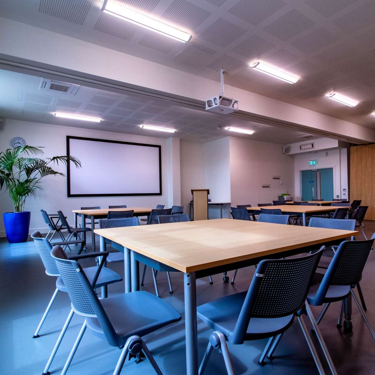A spacious, well-lit meeting room with several wooden tables and chairs arranged around a central area, featuring a large projection screen on one wall.