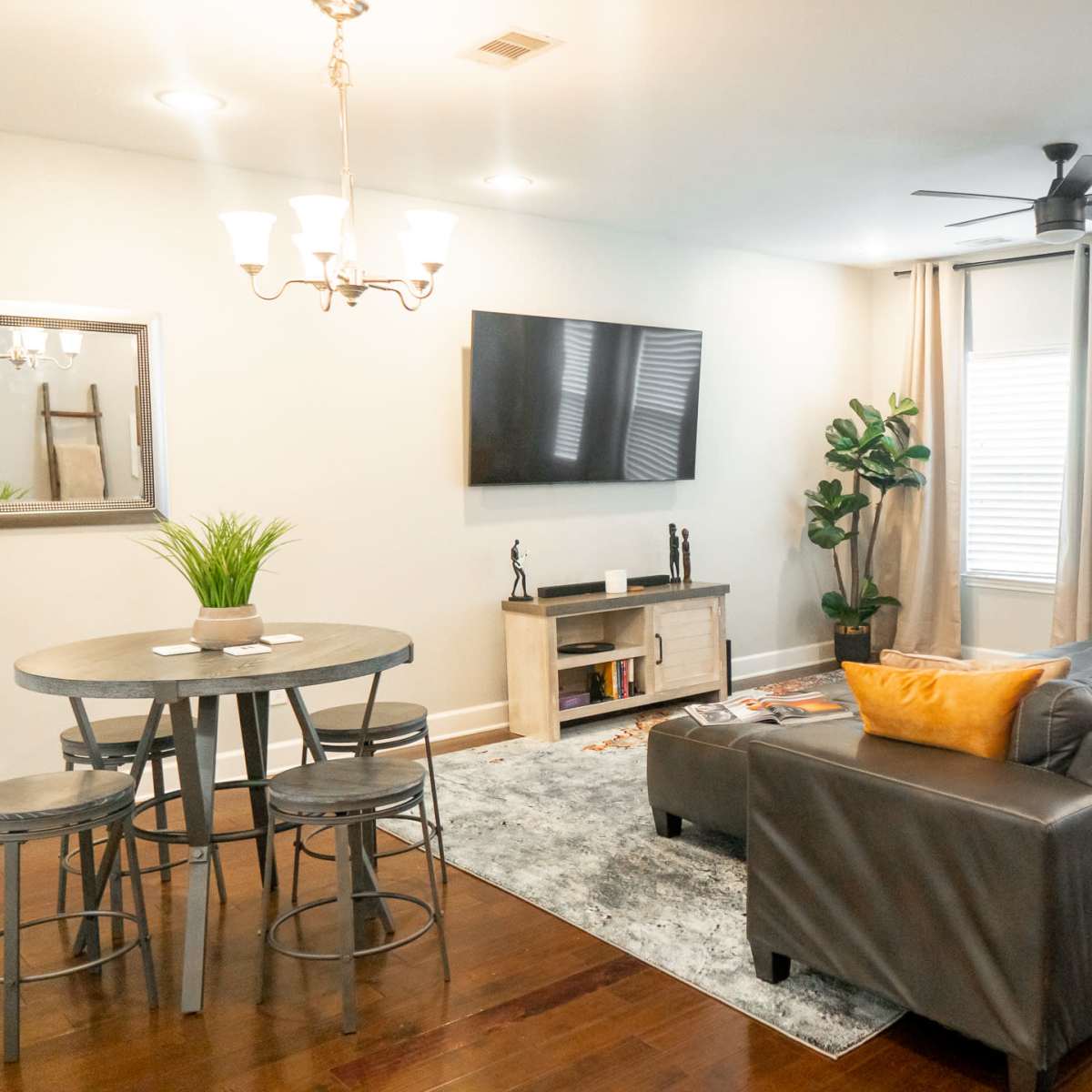 The living room features a gray couch, a round dining table with stools, a wall-mounted television, and large windows with light curtains.