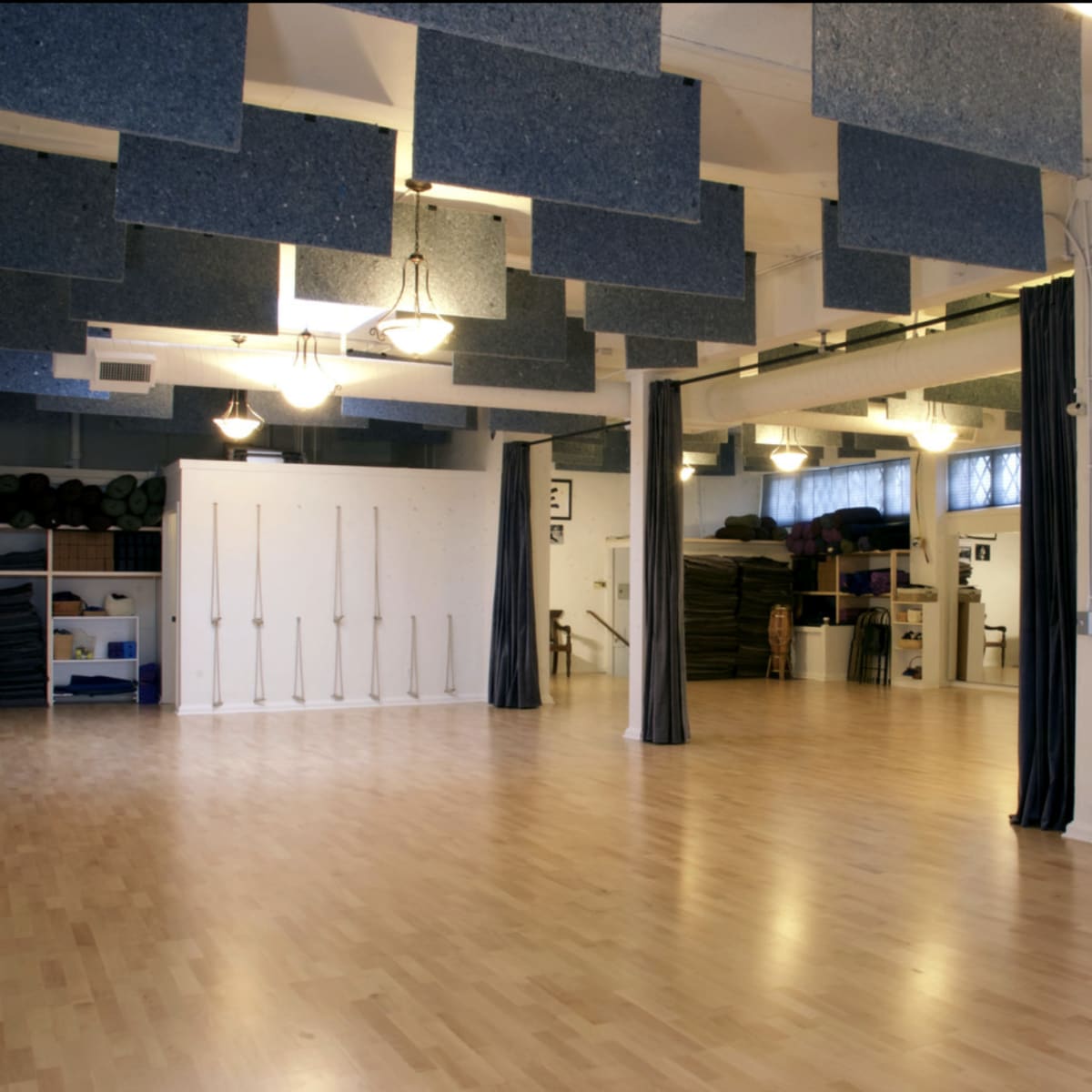 A spacious, well-lit studio with a wooden floor, blue acoustic panels hanging from the ceiling, and curtains dividing different areas.