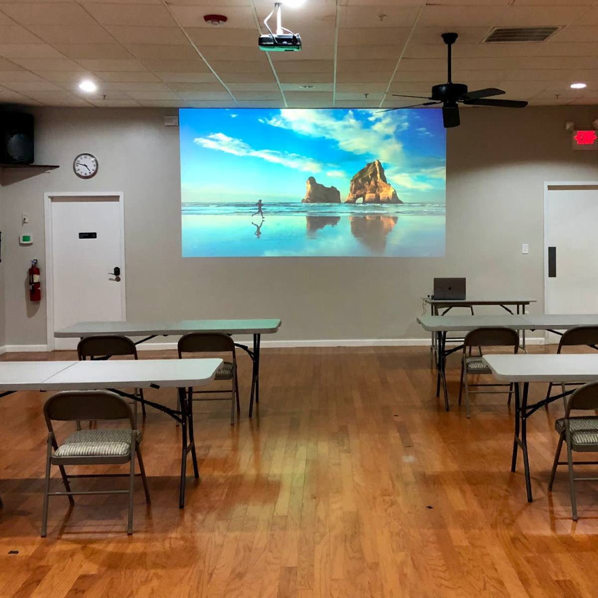 A meeting room features several tables and chairs arranged facing a large projection screen displaying a beach scene.