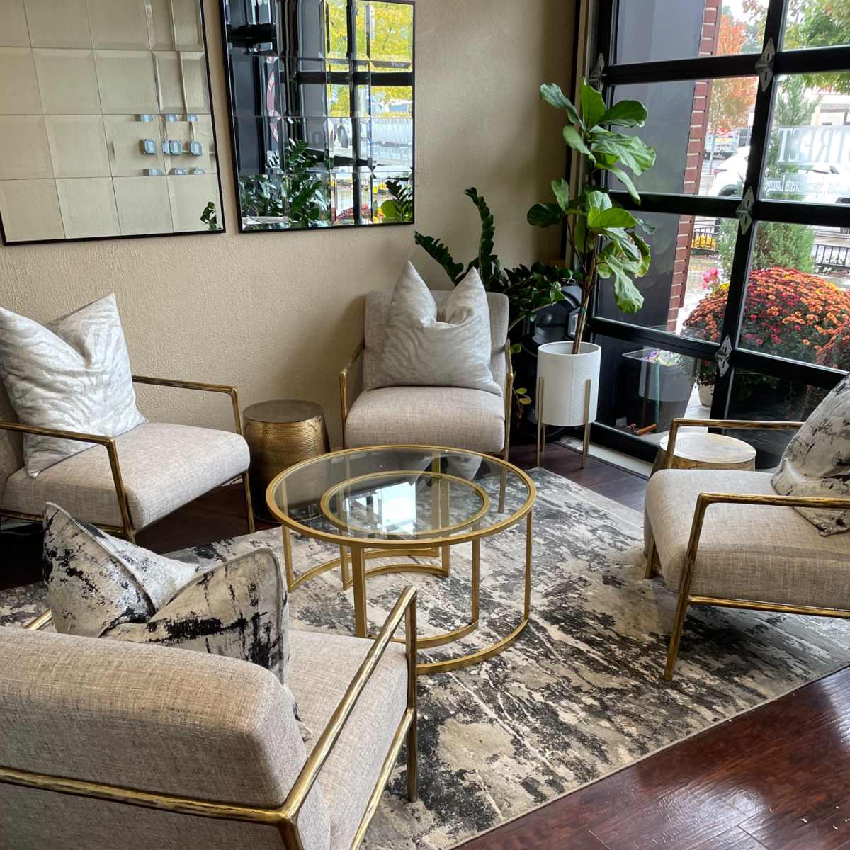 A modern sitting area features four gold-framed chairs arranged around a glass-topped coffee table on a patterned rug, with plants and a mirror in the background.