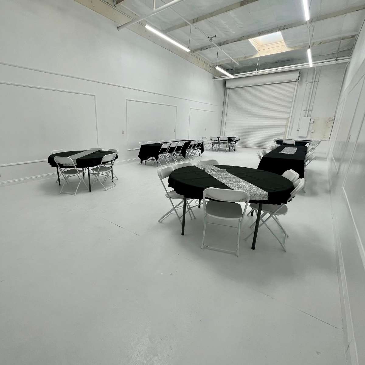 An empty indoor event space with black and white tables arranged in a sparse layout, set against a white background and large roll-up door.