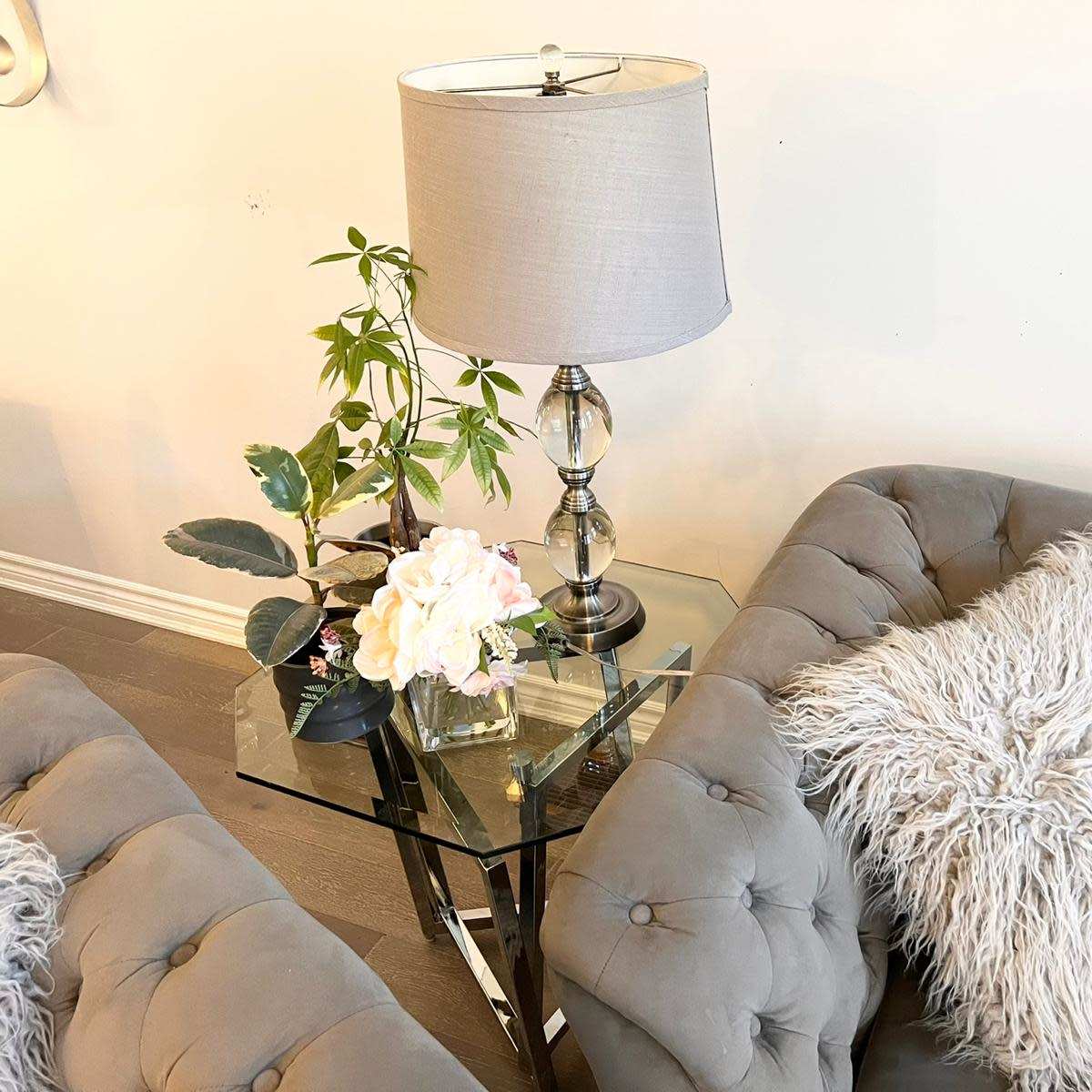 A glass side table with a lamp and a floral arrangement sits between two tufted gray sofas adorned with fluffy pillows.